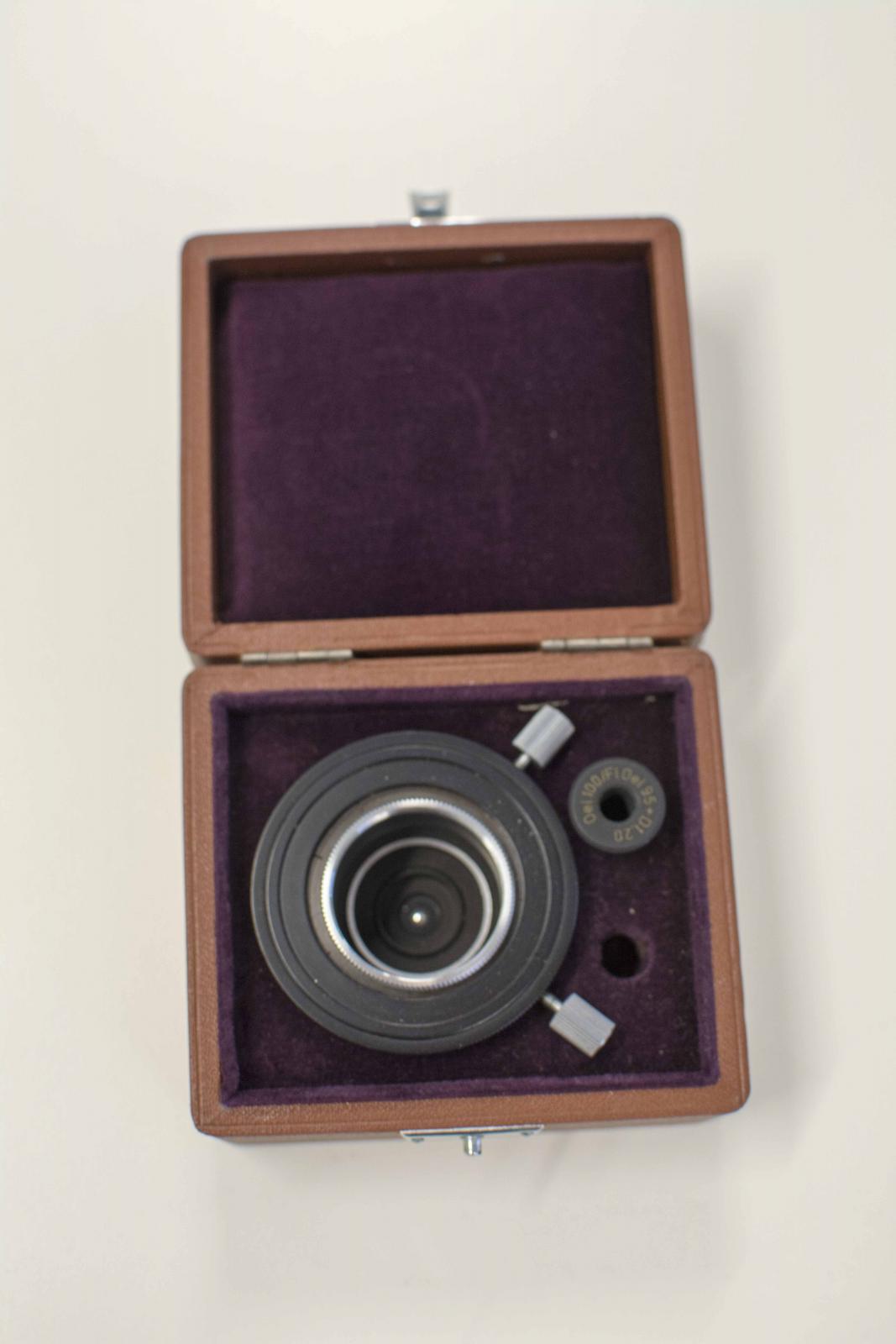 Caja marrón forrada en terciopelo morado en su interior. Contiene una lente negra con botones metálicos
