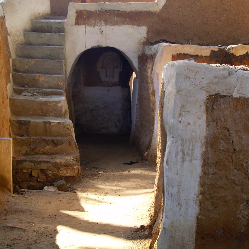 Fotografía. Detalle de un espacio urbano con arquitectura vernácula. A través de una estrecha calle que conduce a un espacio de acceso mediante un arco, la fotografía muestra el entorno urbano de la ciudad de Ghadames