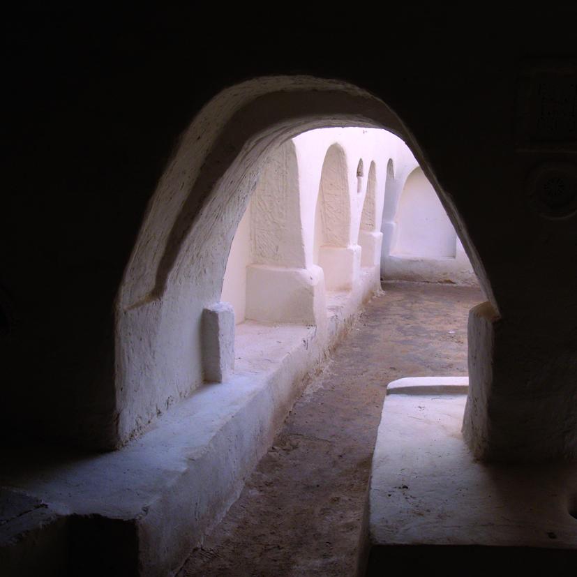 Fotografía de formato cuadrado. Detalle de calle estrecha tomado desde un espacio cubierto y oscuro, abierto al exterior con un arco parabólico que deja ver al otro lado una sucesión de arcadas ciegas con arcos de la misma tipología