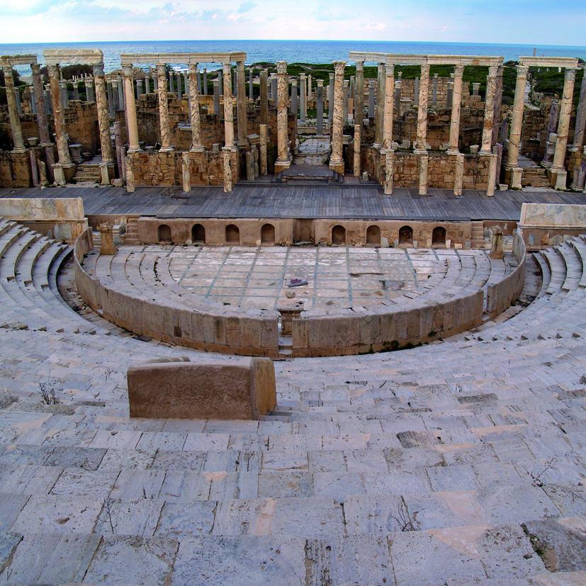 Fotografía de formato cuadrado. Desde la altura del graderío, se observan la orchestra, la escena con hileras de columnas y, al fondo, el mar