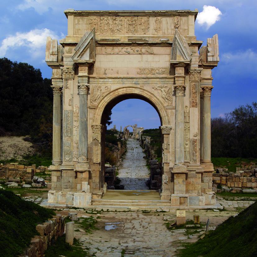 Fotografía de formato cuadrado. Composición centrada por un arco de triunfo de un solo vano a través del que se acentúa la perspectiva, marcada por la línea que dibuja en el suelo una calzada romana.