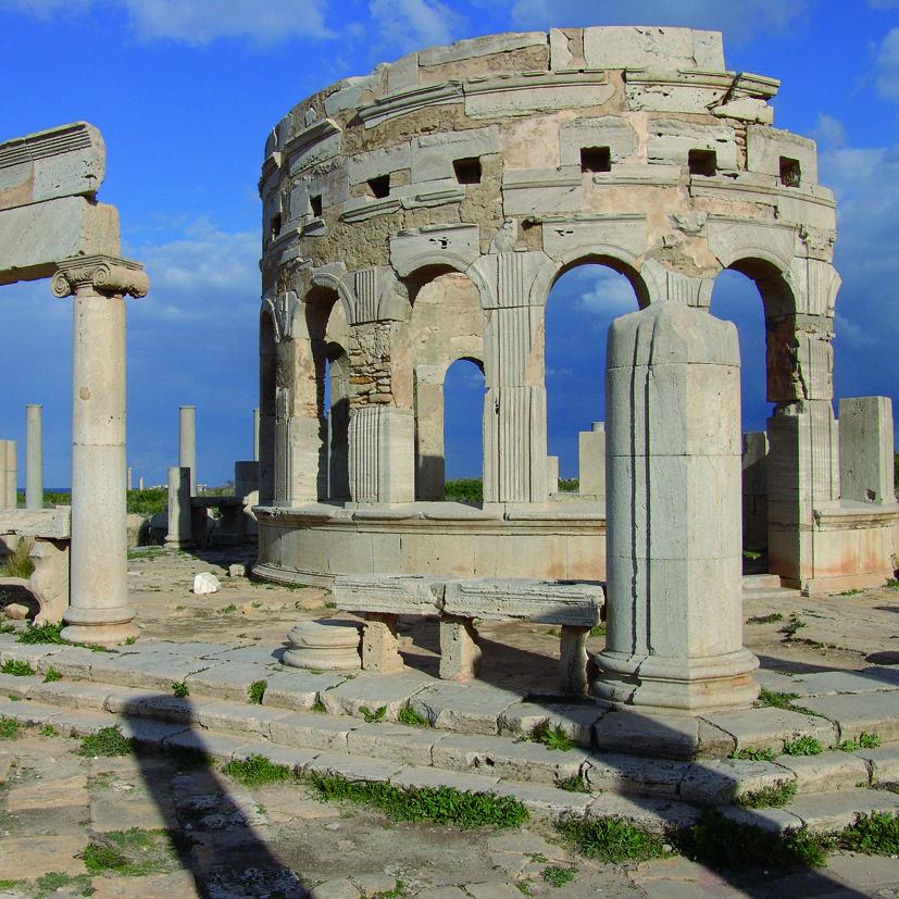 Fotografía de formato cuadrado. Composición que muestra restos arqueológicos romanos entre los que se cuentan columnas y un edificio de planta circular con arcos de medio punto y pilastras acanaladas 