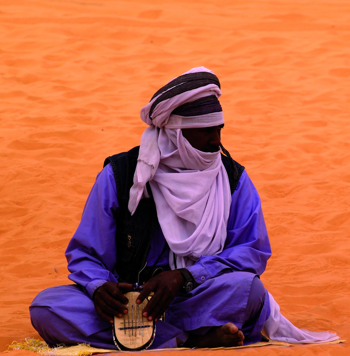 Fotografía de formato cuadrado. En primer plano, un personaje sentado toca un pequeño instrumento, vestido en azules con chilaba, pañuelo turbante lila y chaleco negro. De fondo, la arena del desierto