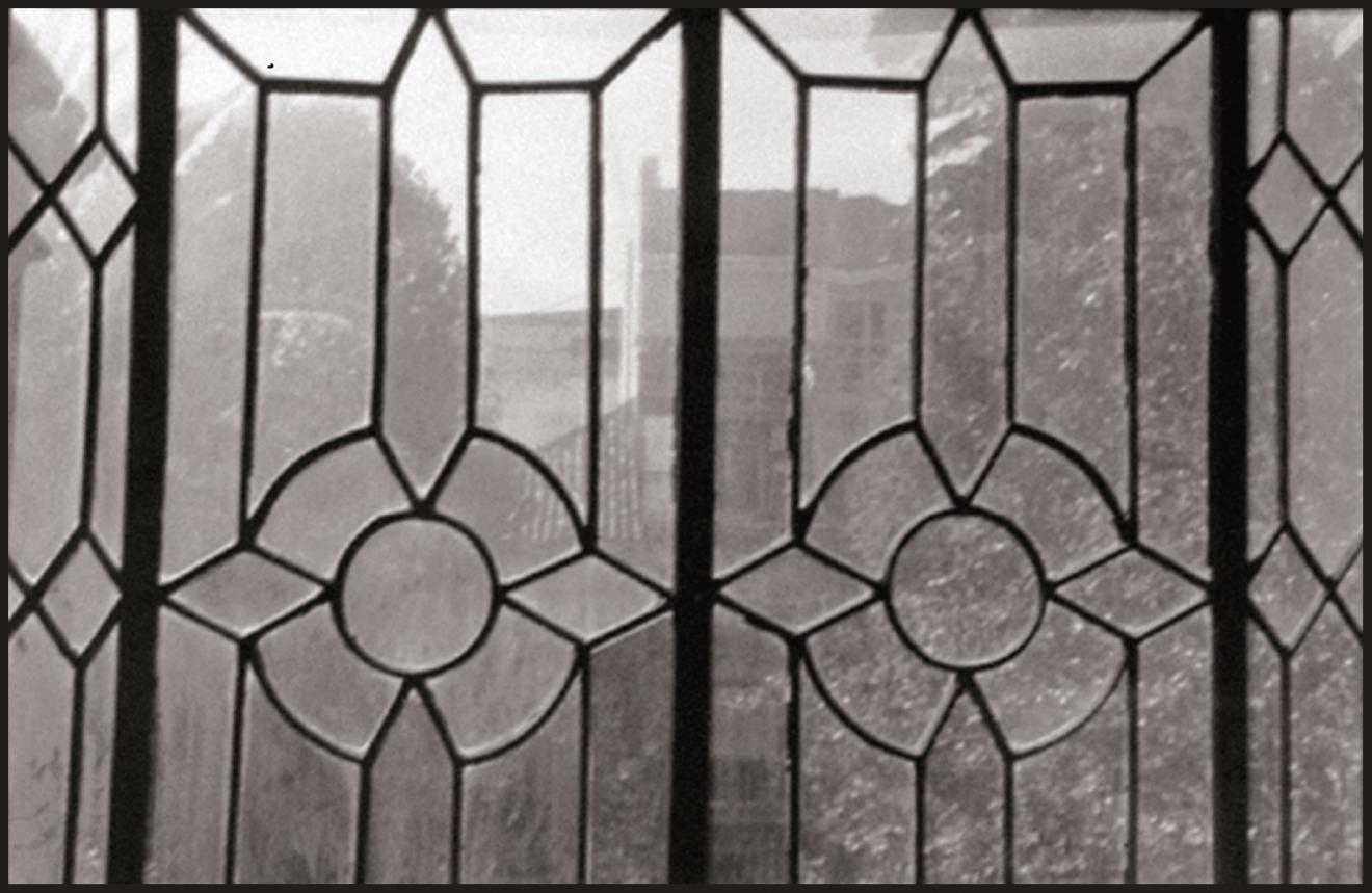 Fotografía en escala de grises de una rejería geometrizante a través de la cual se observa un espacio ajardinado